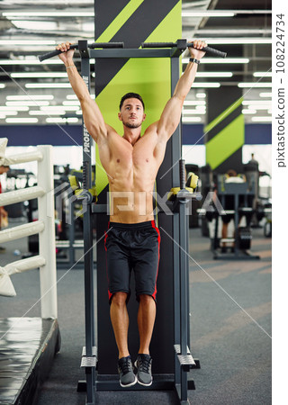 Attractive man does pull-up on the crossbar during training in the modern gym. Front view. Attractive man does pull-up on the crossbar during training in the modern gym. Front view. 108224734