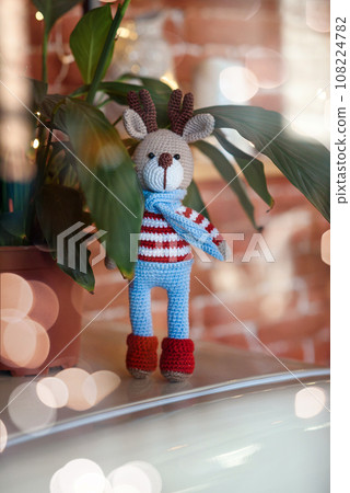 Christmas deer. Closed up toy amigurumi deer in striped sweater and stylish blue scarf stands near flowerpot. 108224782