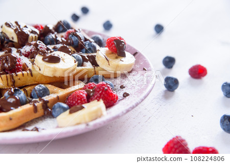 Tasty belgian waffles with blueberries, raspberries, bananas and sugar powder are filled with liquid chocolate. 108224806