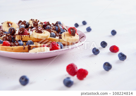 Tasty belgian waffles with blueberries, raspberries, bananas and sugar powder are filled with liquid chocolate. Tasty belgian waffles with blueberries, raspberries, bananas and sugar powder are filled with liquid chocolate. 108224814