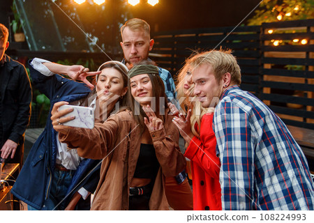 Female and male friends celebrating the party outdoors and making selfies photo on smartphone. Young caucasian people having fun and enjoying weekend at party. 108224993