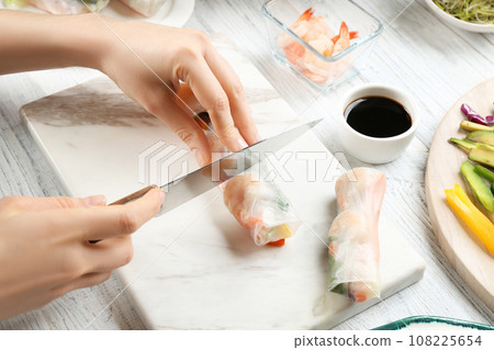 Woman cutting rice paper roll at white wooden table, closeup 108225654