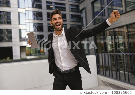 Businessman with laptop standing on office terrace and raised fist is suttisfied of finished project 108225663