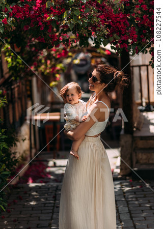 A stylish woman with a baby in her arms on the background of a street in Turkey. A baby in his mother's arms. A beautiful portrait of a child in the arms of his mother. A woman with a baby 108227544