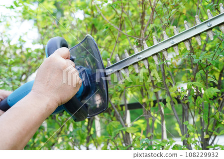 Gardener trimming bush by electric hedge clippers in garden. Hobby at home. Gardener trimming bush by electric hedge clippers in garden. Hobby at home. 108229932