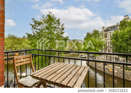 a balcony with a wooden table and two chairs on it, overlooking the canal in the background is a cloudy blue sky a balcony with a wooden table and two chairs on it, overlooking the canal in the background is a cloudy blue sky 108231822