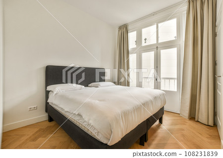 a bed in a room with wood flooring and curtains on either side of the bed are two large windows a bed in a room with wood flooring and curtains on either side of the bed are two large windows 108231839