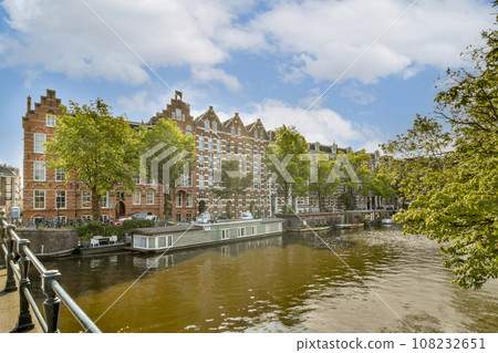 some buildings on the side of a body of water with trees in the foreground and people walking along it 108232651