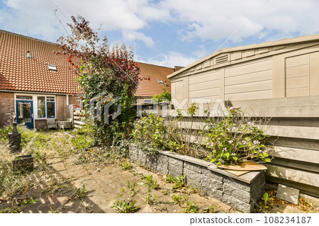 an outside area with some plants and flowers on the ground in front of a house, next to a brick wall an outside area with some plants and flowers on the ground in front of a house, next to a brick wall 108234187
