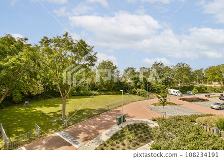 a park with cars parked in the parking lot and people walking on the sidewalk to the right, there is a cloudy blue sky 108234191