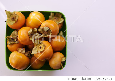 Bunch of ripe persimmons lies in a square green bowl on a white table. Top view Bunch of ripe persimmons lies in a square green bowl on a white table. Top view 108237894