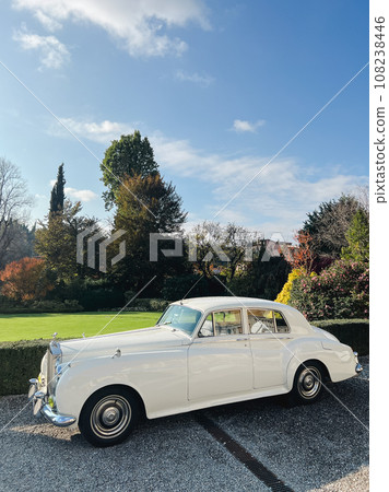 White Rolls-Royce Silver Cloud stands on a gravel path near green bushes in the park White Rolls-Royce Silver Cloud stands on a gravel path near green bushes in the park 108238446