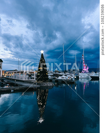 Christmas tree decorated with luminous garlands stands in the port near the yachts 108239086