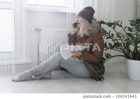 A woman warmly dressed in a brown jacket and hat is sitting near the heater with bills in her hands and shouting on the phone. The concept of a crisis and a large set for utilities. 108241643