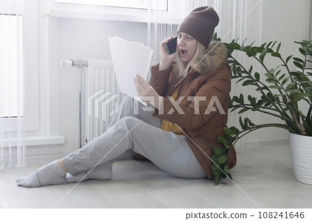 A woman warmly dressed in a brown jacket and hat is sitting near the heater with bills in her hands and shouting on the phone. The concept of a crisis and a large set for utilities. 108241646