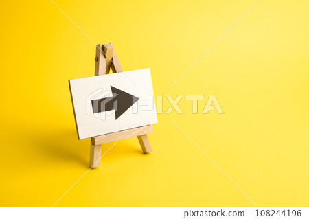 Arrow on easel. Advertising sign. Direction pointer. Hint, help in navigation. Minimalism. Advertising and attracting attention. Redirecting traffic. Short road. Point at, show the way. Arrow on easel. Advertising sign. Direction pointer. Hint, help in navigation. Minimalism. Advertising and attracting attention. Redirecting traffic. Short road. Point at, show the way. 108244196