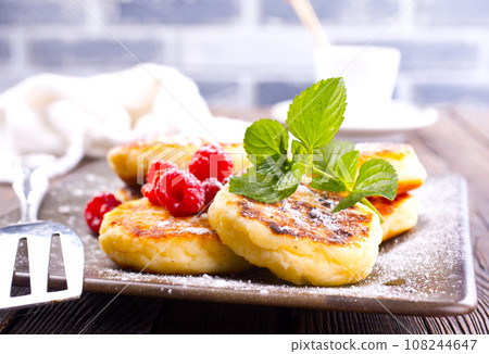 Traditional sweet syrniki or cottage cheese pancakes served with sweet cream and fresh raspberries in a white plate over table. Traditional sweet syrniki or cottage cheese pancakes served with sweet cream and fresh raspberries in a white plate over table. 108244647