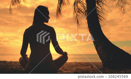 Woman meditate sitting on beach under palm tree against bright orange sunset sky. Girl silhouette in lotus pose enjoys beautiful nature landscape. Tropical vacation concept. Travel, tourism, holiday 108245569