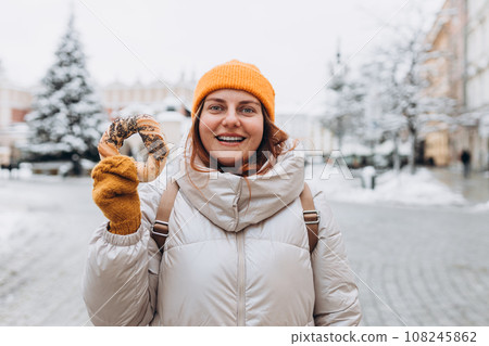 Attractive young female tourist in yellow hat is holding pretzel, traditional polish bagel on Market Square in Krakow. Traveling Europe in winter time. High quality photo Attractive young female tourist in yellow hat is holding pretzel, traditional polish bagel on Market Square in Krakow. Traveling Europe in winter time. High quality photo 108245862