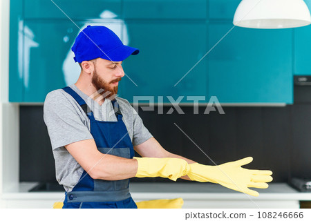 Attractive guy with well-groomed beard from cleaning service wearing protective rubber gloves to clean the domestic cuisine Attractive guy with well-groomed beard from cleaning service wearing protective rubber gloves to clean the domestic cuisine 108246666