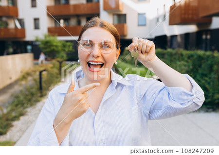 Attractive young woman showing key while standing outdoor against new house. Real estate agent holding key on street background 108247209