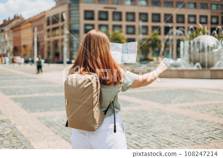 Attractive young female tourist is exploring new city. Redhead woman pointing finger and holding a paper map in a warm sunny day. Urban lifestyle concept. Traveler 108247214