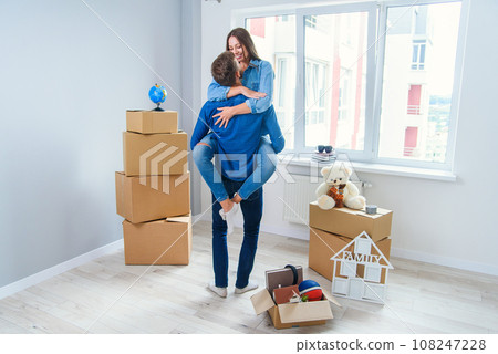 Front view of pretty smiling girl which holds by her handsome young boyfriend in the middle of their new flat full of cardboard boxes. Front view of pretty smiling girl which holds by her handsome young boyfriend in the middle of their new flat full of cardboard boxes. 108247228