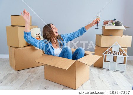 Happy woman drinks champagne from a glass while sitting inside a cardboard box in new apartment. Happy woman drinks champagne from a glass while sitting inside a cardboard box in new apartment. 108247237