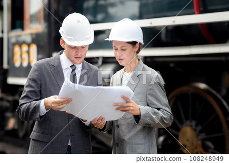 Young caucasian engineer man and woman in suit checking train looking blueprint in station. 108248949