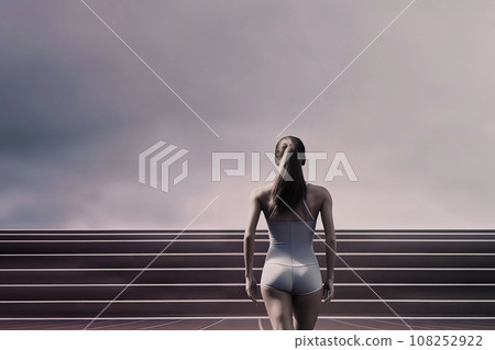 Surreal minimalist rear view of a female athlete in skimpy sportswear in front of a running track with the gray sky in the background, copy space 108252922