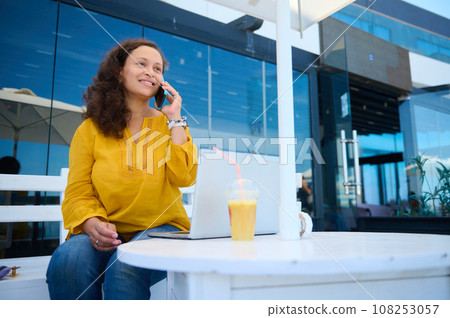 Beautiful multi-ethnic middle aged young woman sitting at a cafe with open laptop, smiling while talking on mobile phone. People. Online business and remote work concept 108253057