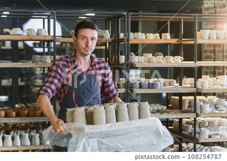 Caucasian male potter holding tray with ceramic pots in the pottery. Caucasian male potter holding tray with ceramic pots in the pottery. 108254787
