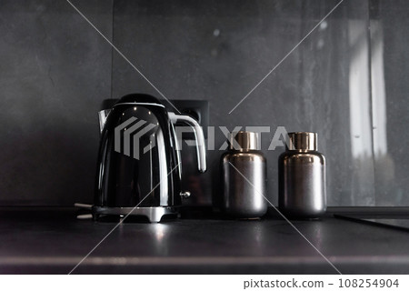 Modern black kettle and bottles for spices standing on black kitchen workspace on the background of grey tile like a marble. Modern black kettle and bottles for spices standing on black kitchen workspace on the background of grey tile like a marble. 108254904