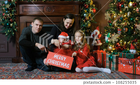 Cheerful family having fun near Xmas tree, pointing fingers on sign with inscription Merry Christmas. Winter holidays and New Year concept 108255035