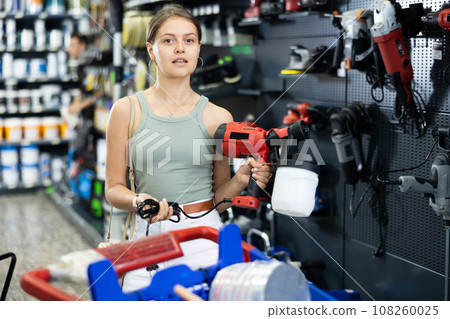 Beautiful girl planning home renovation and choosing paint sprayer in hardware store Beautiful girl planning home renovation and choosing paint sprayer in hardware store 108260025