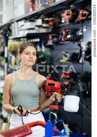 Female buyer doubts choice and closely examines electric paint-spraying machine in supermarket. Repair and construction items Female buyer doubts choice and closely examines electric paint-spraying machine in supermarket. Repair and construction items 108260054