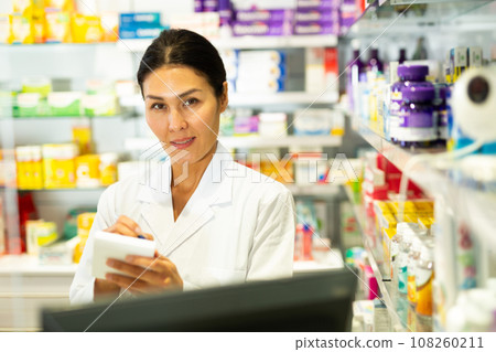 Medicine, profession and healthcare concept - female doctor or pharmacist in white coat with notebook and pen writing medical report over pharmacy Medicine, profession and healthcare concept - female doctor or pharmacist in white coat with notebook and pen writing medical report over pharmacy 108260211