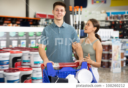 Spouses shopper stand with trolley filled with paint goods for repair and coloring in store sales hall. Spouses shopper stand with trolley filled with paint goods for repair and coloring in store sales hall. 108260212