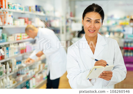 Portrait of female pharmacist working in pharmacy during the pandemic, standing in trading floor and makes important notes Portrait of female pharmacist working in pharmacy during the pandemic, standing in trading floor and makes important notes 108260546