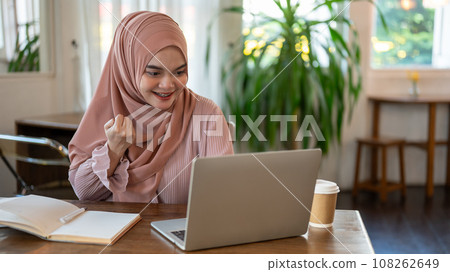 A cheerful Asian-Muslim woman is looking at her laptop screen with a happy face, good news. 108262649