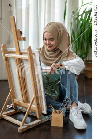 Happy Asian-Muslim female artist is sitting on the floor, enjoying creating her artwork on a canvas 108262719