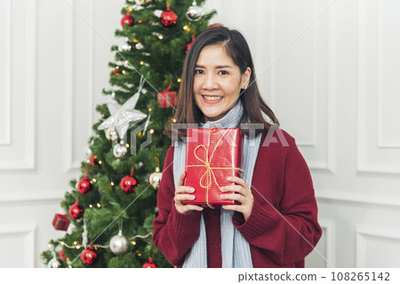 Woman surprise got present gift box on christmas eve. Asian woman holding christmas gift box under xmas tree with smiling face. Woman hands wrap present gift. Merry christmas happy new year concept 108265142