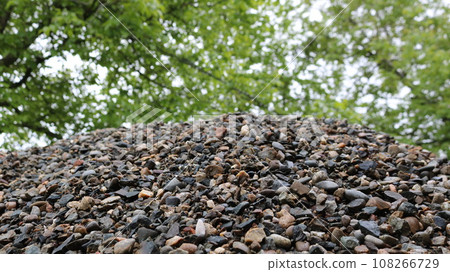 bottom view of a mountain of gray gravel on a blurred background of green tree foliage, landscape details with small rock lying in a crumbly pile, a fragment of a stone embankment as a natural texture 108266729