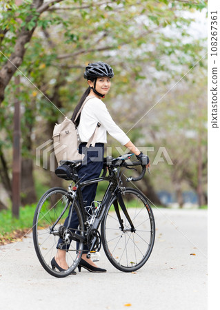 A young woman walking while pushing a road bike in business casual clothes, back view 108267361