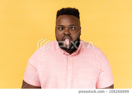 Portrait of disobedient crazy funny man wearing pink shirt showing tongue out and crossed his eyes displeased, behaving unruly naughty. Indoor studio shot isolated on yellow background. Portrait of disobedient crazy funny man wearing pink shirt showing tongue out and crossed his eyes displeased, behaving unruly naughty. Indoor studio shot isolated on yellow background. 108268899