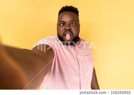 Portrait of funny childish bearded man wearing pink shirt making point of view photo, looking at camera with crossed eyes and tongue out. Indoor studio shot isolated on yellow background. Portrait of funny childish bearded man wearing pink shirt making point of view photo, looking at camera with crossed eyes and tongue out. Indoor studio shot isolated on yellow background. 108268902
