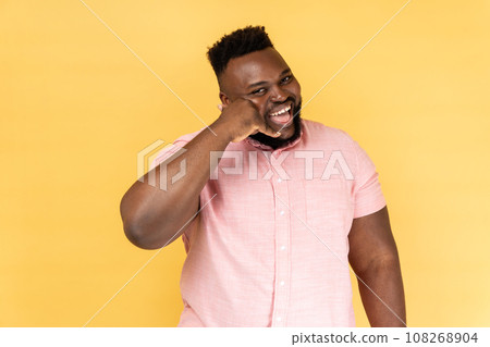 Portrait of satisfied man wearing pink shirt flirting holding fingers near ear showing call gesture with toothy smile, answering call. Indoor studio shot isolated on yellow background. Portrait of satisfied man wearing pink shirt flirting holding fingers near ear showing call gesture with toothy smile, answering call. Indoor studio shot isolated on yellow background. 108268904