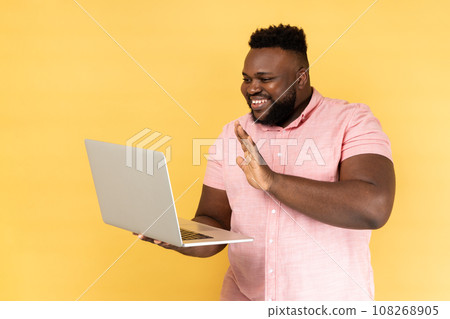 Portrait of friendly man wearing pink shirt waving hand, gesturing hello to laptop screen, video call with friend, communicating online. Indoor studio shot isolated on yellow background. Portrait of friendly man wearing pink shirt waving hand, gesturing hello to laptop screen, video call with friend, communicating online. Indoor studio shot isolated on yellow background. 108268905