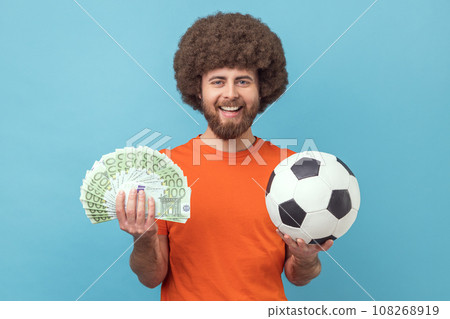 Portrait of happy smiling man with Afro hairstyle wearing orange T-shirt holding soccer ball and fan of euro bills, sports betting, big win. Indoor studio shot isolated on blue background. Portrait of happy smiling man with Afro hairstyle wearing orange T-shirt holding soccer ball and fan of euro bills, sports betting, big win. Indoor studio shot isolated on blue background. 108268919