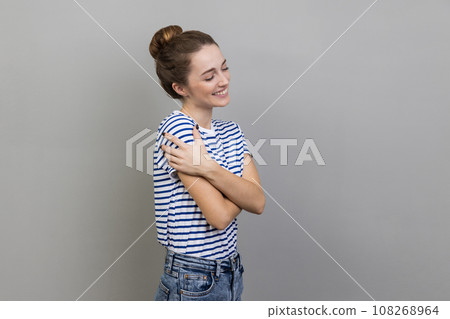 Side view portrait of cheerful woman wearing striped T-shirt smiles tenderly, touches shoulders, embraces herself, keeps eyes closed. Indoor studio shot isolated on gray background. 108268964